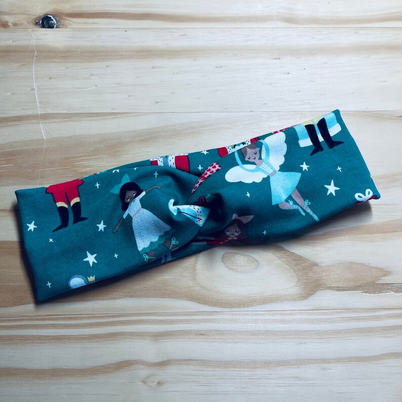 May include: A teal blue fabric headband with a Christmas-themed print featuring a nutcracker, a ballerina, and stars. The headband is twisted and tied in a knot.