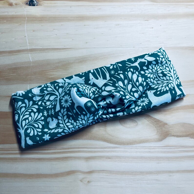 May include: A green and white patterned headband with a knotted design. The pattern features trees, birds, and deer.