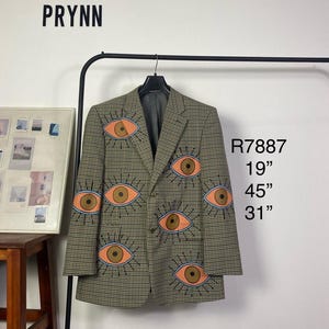 May include: A tan and black patterned blazer adorned with eye appliqués in orange and blue. The text "PRYNN" is visible. Measurements are 48cm, 114cm, and 79cm.