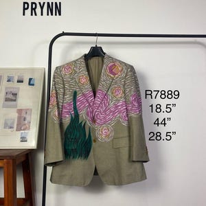May include: A tan blazer with a colorful, artistic design. The blazer features a pink and white swirl pattern on the upper portion and green flame-like shapes. Measurements are 47 cm chest, 112 cm length, and 72 cm sleeve.