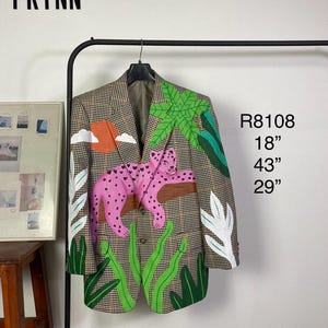 May include: A brown and black houndstooth blazer featuring a hand-painted design of a pink leopard resting on a branch, surrounded by green palm leaves, white foliage, and an orange sun. Text includes PRYNN, R8108, 18", 43", and 29".