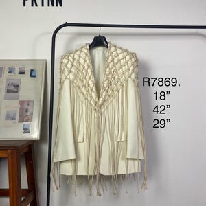 Puede incluir: Blazer color crema con un diseño de hombros tejido único y detalles de flecos. La chaqueta tiene cuello en V y dos bolsillos. Medidas: 45.7 cm de ancho, 106.7 cm de largo y 73.7 cm de largo de manga. La palabra "PRYNN" está arriba.