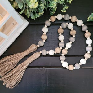 May include: A decorative wooden bead garland with tassels. The garland features a mix of white, gray, and tan wooden beads. Two jute tassels hang from the end of the garland. The garland is displayed on a black surface with greenery in the background.