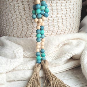 May include: A wooden bead garland with teal and natural wood beads and two brown tassels. The garland is hanging from a woven basket.
