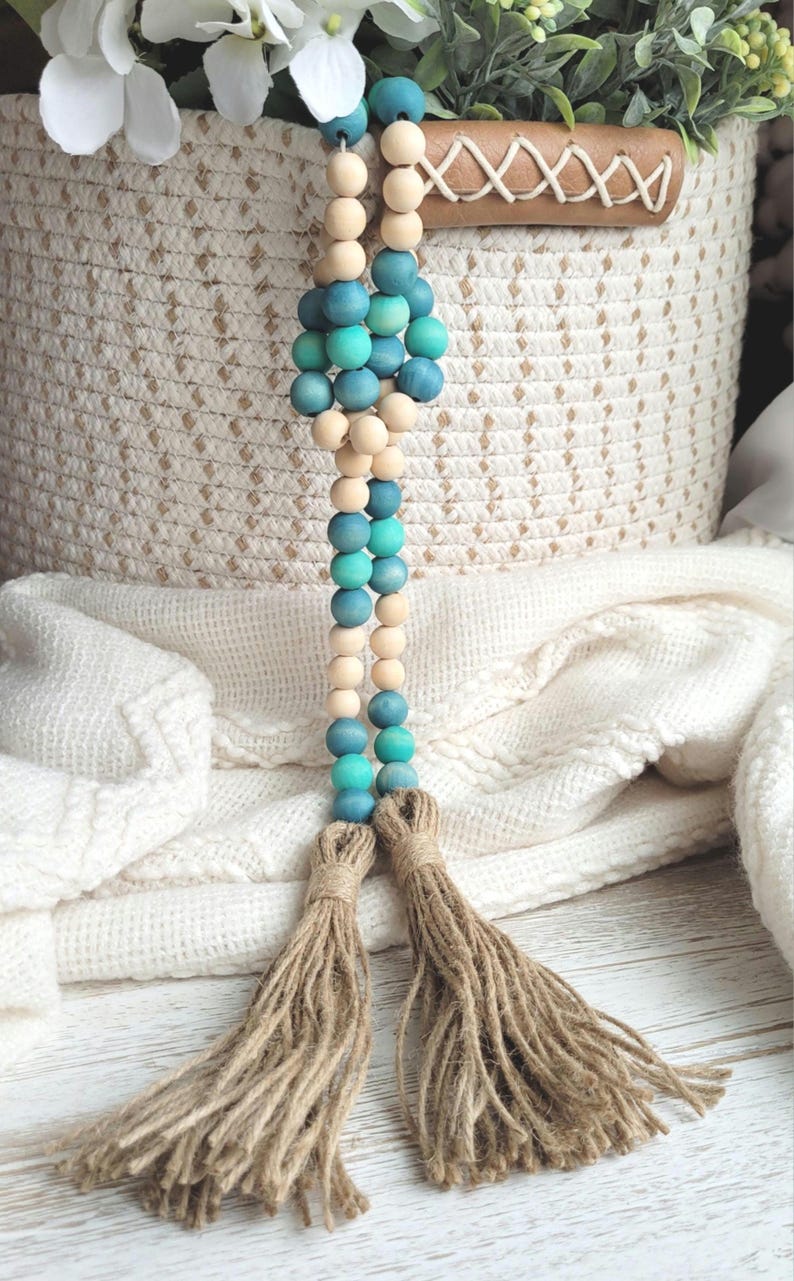 May include: A wooden bead garland with teal and natural wood beads and two brown tassels. The garland is hanging from a woven basket.