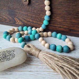 May include: A teal and natural wood bead garland with a jute tassel. The garland is draped over a wooden box and a white stone with the word "Relax" written on it.