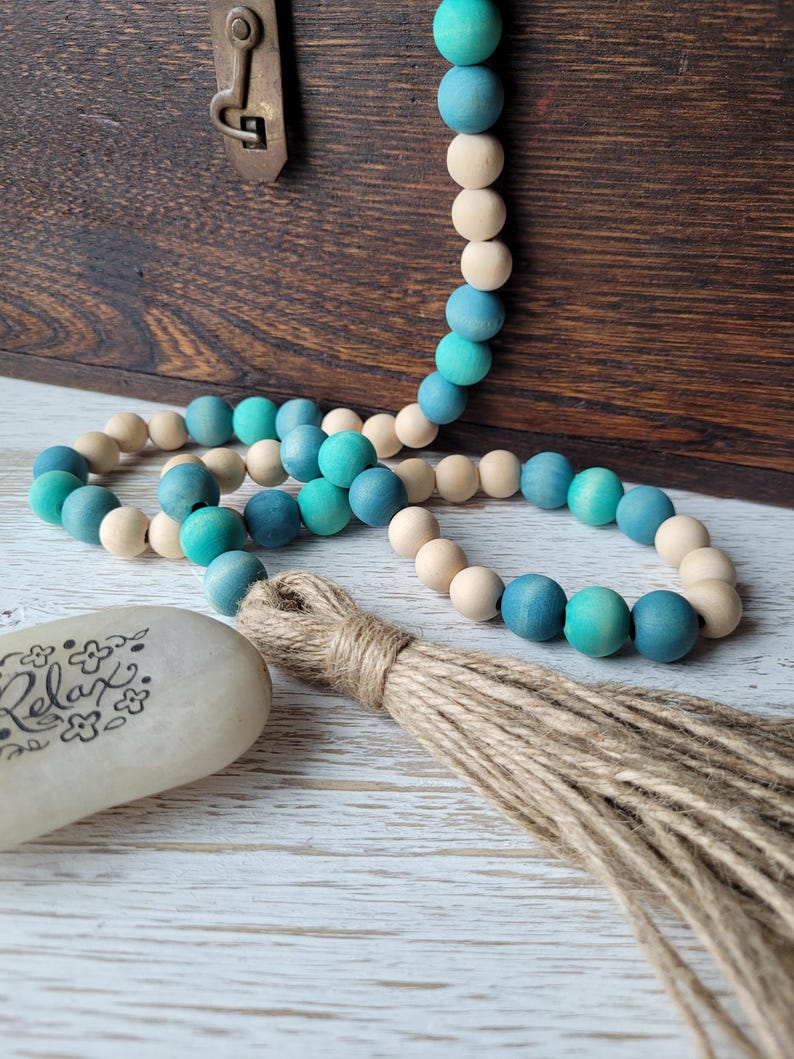 May include: A teal and natural wood bead garland with a jute tassel. The garland is draped over a wooden box and a white stone with the word "Relax" written on it.