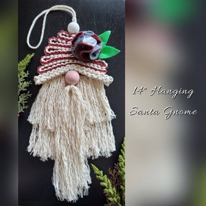 May include: A 14-inch hanging Santa gnome decoration. The gnome features a cream-colored beard and a red and cream striped hat. A pink wooden nose and a fabric flower with green leaves adorn the hat. The text "14" Hanging Santa Gnome" is visible.