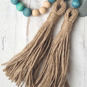 May include: Two brown jute tassels with a natural fibre texture. The tassels are attached to a string of wooden beads with a blue ombre finish.
