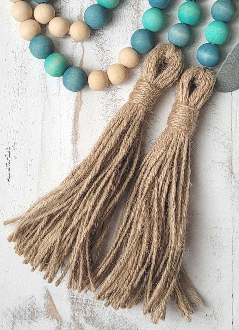 May include: Two brown jute tassels with a natural fibre texture. The tassels are attached to a string of wooden beads with a blue ombre finish.