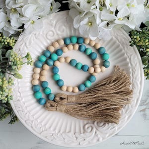 May include: A wooden bead garland with a gradient of teal and blue colours. The garland has two brown tassels at the end. The garland is on a white plate with a floral design.