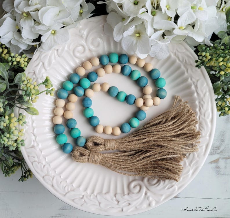 May include: A wooden bead garland with a gradient of teal and blue colours. The garland has two brown tassels at the end. The garland is on a white plate with a floral design.