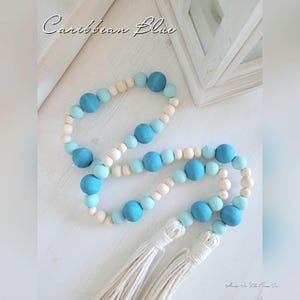 May include: Decorative wooden bead garland in shades of Caribbean blue and natural wood tones. The garland features large and small round beads, with two white tassel accents. The text "Caribbean Blue" is written in cursive at the top.