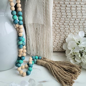 May include: A wooden bead garland with an ombre effect in shades of teal and natural wood. The garland has a jute tassel at the end.