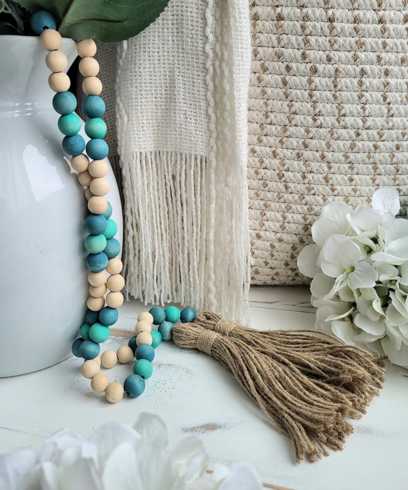 May include: A wooden bead garland with an ombre effect in shades of teal and natural wood. The garland has a jute tassel at the end.
