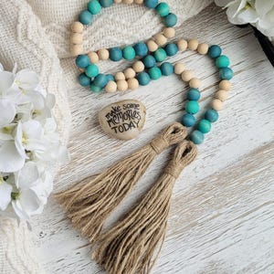 May include: A teal and natural wood bead garland with two brown tassels. The garland has a heart-shaped stone with the text "make some memories today" on it.