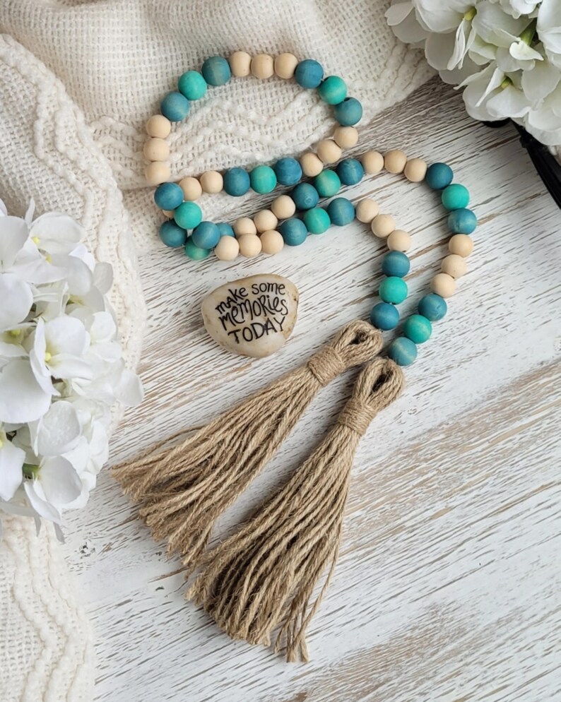 May include: A teal and natural wood bead garland with two brown tassels. The garland has a heart-shaped stone with the text "make some memories today" on it.