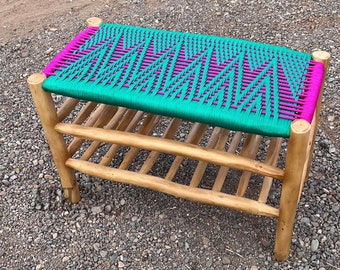 May include: A wooden bench with a woven seat made of green and pink rope. The bench has three shelves and is sitting on a gravel surface.