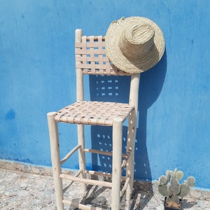 Könnte beinhalten: Ein Barhocker aus Holz mit einem geflochtenen Ledersitz und -lehne. Der Hocker ist hellbraun und hat einen rustikalen Look. Ein Strohhut lehnt an dem Hocker. Der Hocker steht auf einem gefliesten Boden.