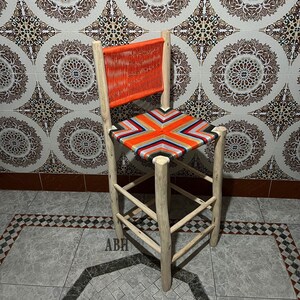 Peut inclure: Un tabouret en bois avec un siège tissé en rayures orange, blanches et noires. Le tabouret a un design simple avec quatre pieds et un siège rectangulaire. Le tabouret est devant un mur carrelé avec un motif marocain.