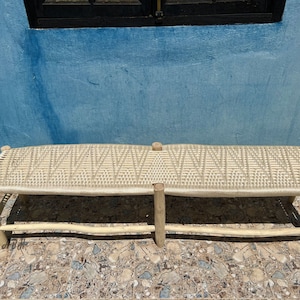 May include: A long, woven bench with a natural wood frame. The bench is made of woven rope and has a light brown color. The bench is sitting on a tiled floor.