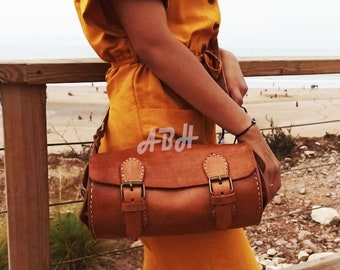 May include: A woman wearing a yellow jumpsuit is standing on a wooden deck overlooking a beach. She is carrying a brown leather satchel with two buckles and the text "ABH" stitched on the front.
