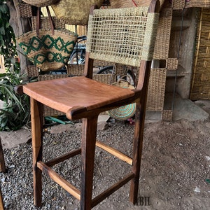 May include: A wooden bar stool with a woven straw back and a brown leather seat. The stool is in front of a wall with woven baskets.