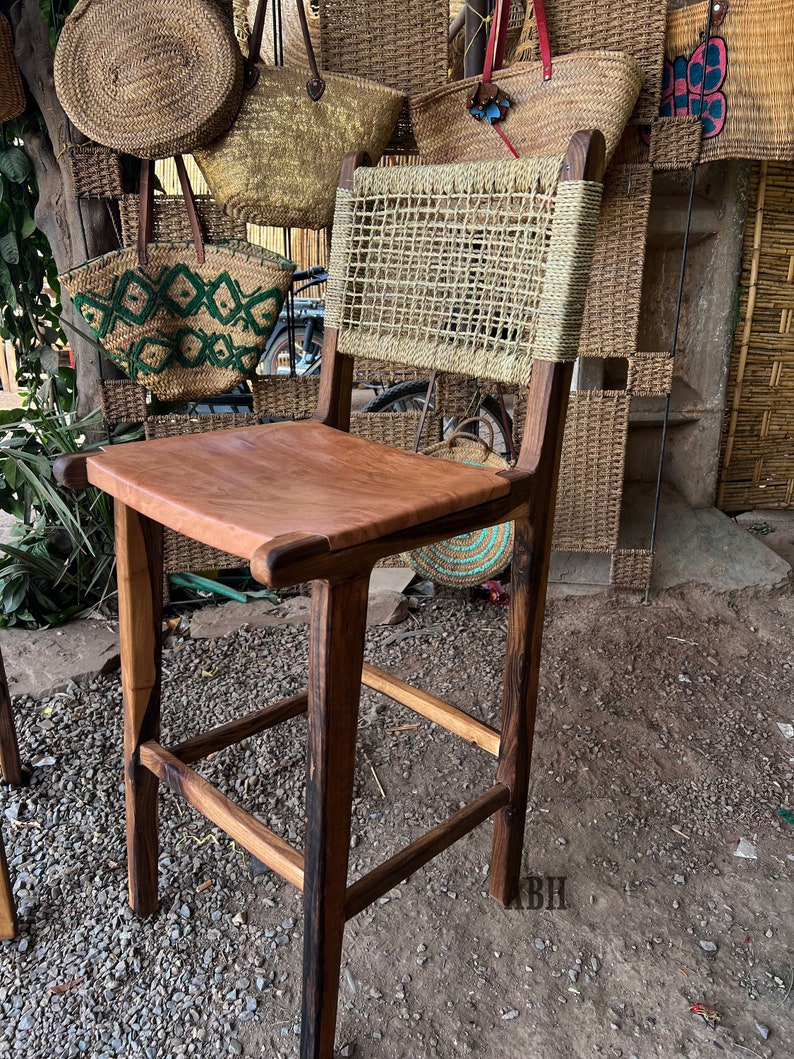May include: A wooden bar stool with a woven straw back and a brown leather seat. The stool is in front of a wall with woven baskets.