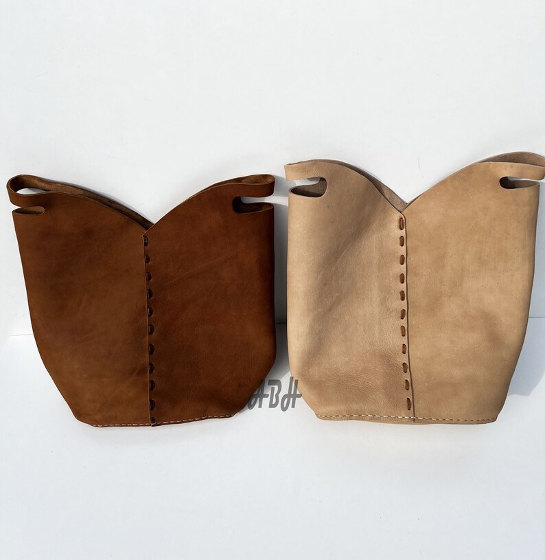 May include: Two leather tote bags, one brown and one beige, with stitched detailing. The bags have a wide, open top and a slightly curved shape.
