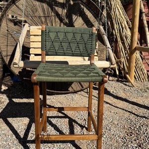 Op de afbeelding: Een houten barstoel met een groene geweven zitting en rugleuning. De zitting en rugleuning zijn geweven met een geometrisch patroon van driehoeken.