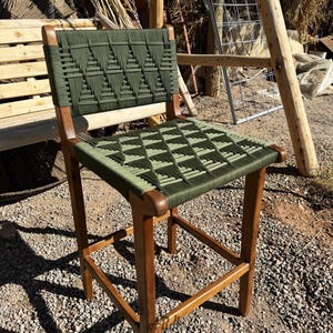 Op de afbeelding: Een houten barstoel met een groene geweven zitting en rugleuning. De stoel heeft een eenvoudig ontwerp met vier poten en een vierkante basis.
