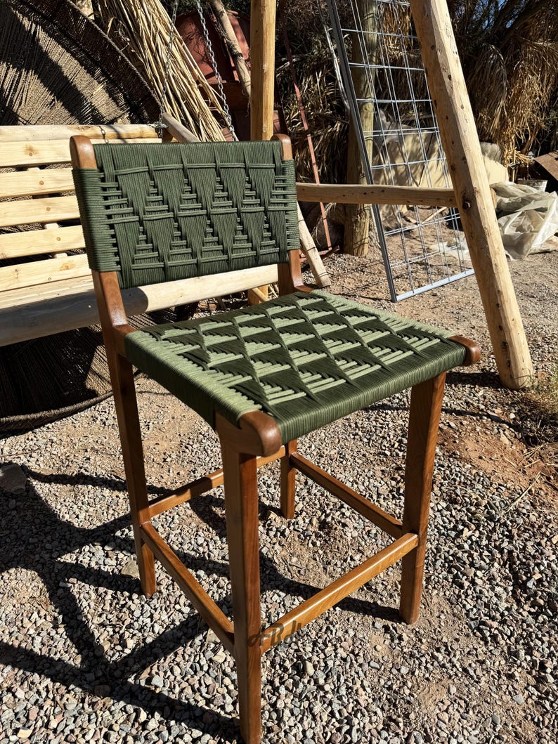 Op de afbeelding: Een houten barstoel met een groene geweven zitting en rugleuning. De stoel heeft een eenvoudig ontwerp met vier poten en een vierkante basis.