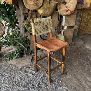 May include: A wooden bar stool with a woven back and a natural wood seat. The stool is sitting on a gravel surface in front of a wall of woven baskets.