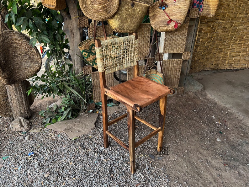 May include: A wooden bar stool with a woven back and a natural wood seat. The stool is sitting on a gravel surface in front of a wall of woven baskets.