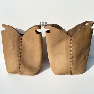 May include: Two tan leather storage baskets with stitched detailing. The baskets are empty and have a slightly distressed look.