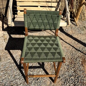 Op de afbeelding: Een groene geweven stoel met een houten frame. De stoel heeft een geometrisch patroon en staat op een grind oppervlak.