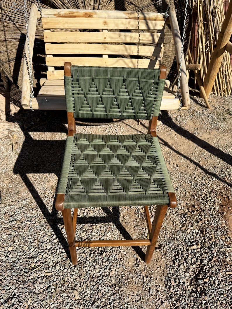 Op de afbeelding: Een groene geweven stoel met een houten frame. De stoel heeft een geometrisch patroon en staat op een grind oppervlak.