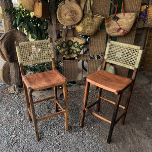May include: Two wooden bar stools with woven cane seats and brown leather cushions. The stools are in front of a wall with woven baskets and bags.