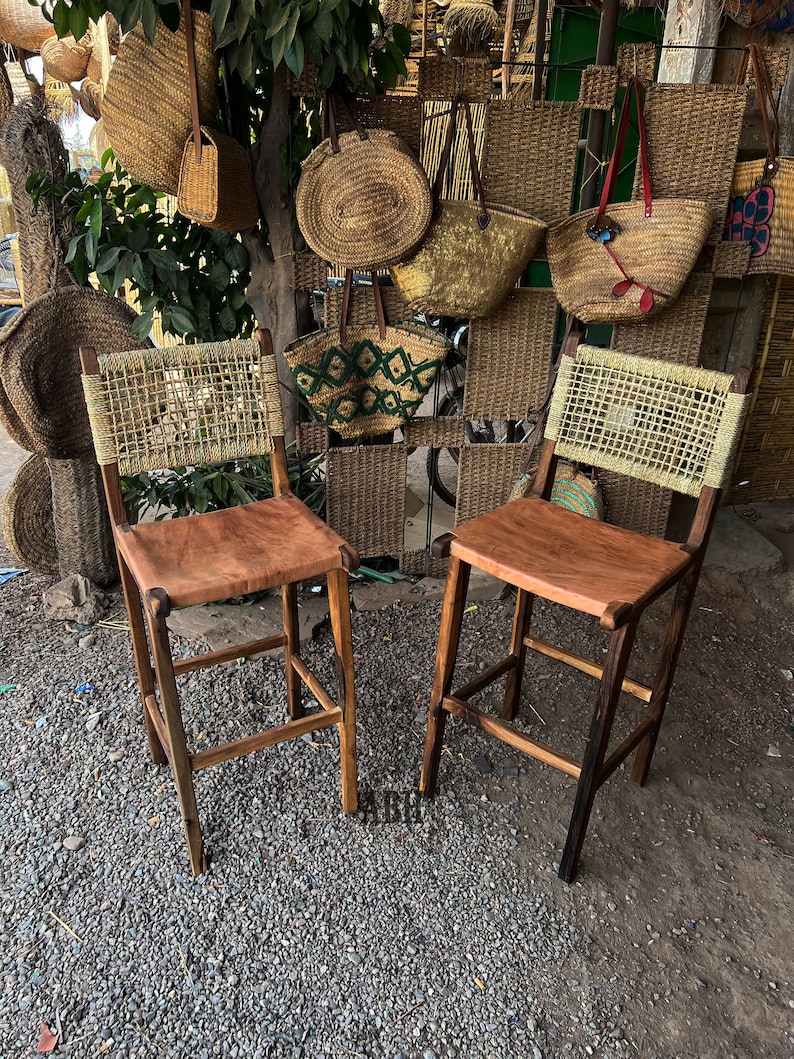 May include: Two wooden bar stools with woven cane seats and brown leather cushions. The stools are in front of a wall with woven baskets and bags.