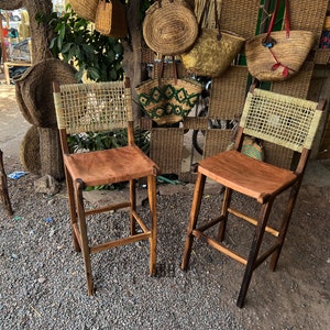 May include: Two wooden bar stools with woven cane backs and brown leather seats. The stools are in front of a wall with woven baskets and bags.