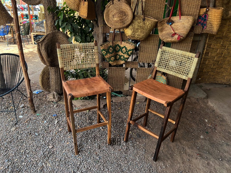 May include: Two wooden bar stools with woven cane backs and brown leather seats. The stools are in front of a wall with woven baskets and bags.