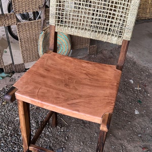 May include: A wooden chair with a woven back and a brown leather seat. The chair has a dark brown finish and is sitting on a gravel surface.