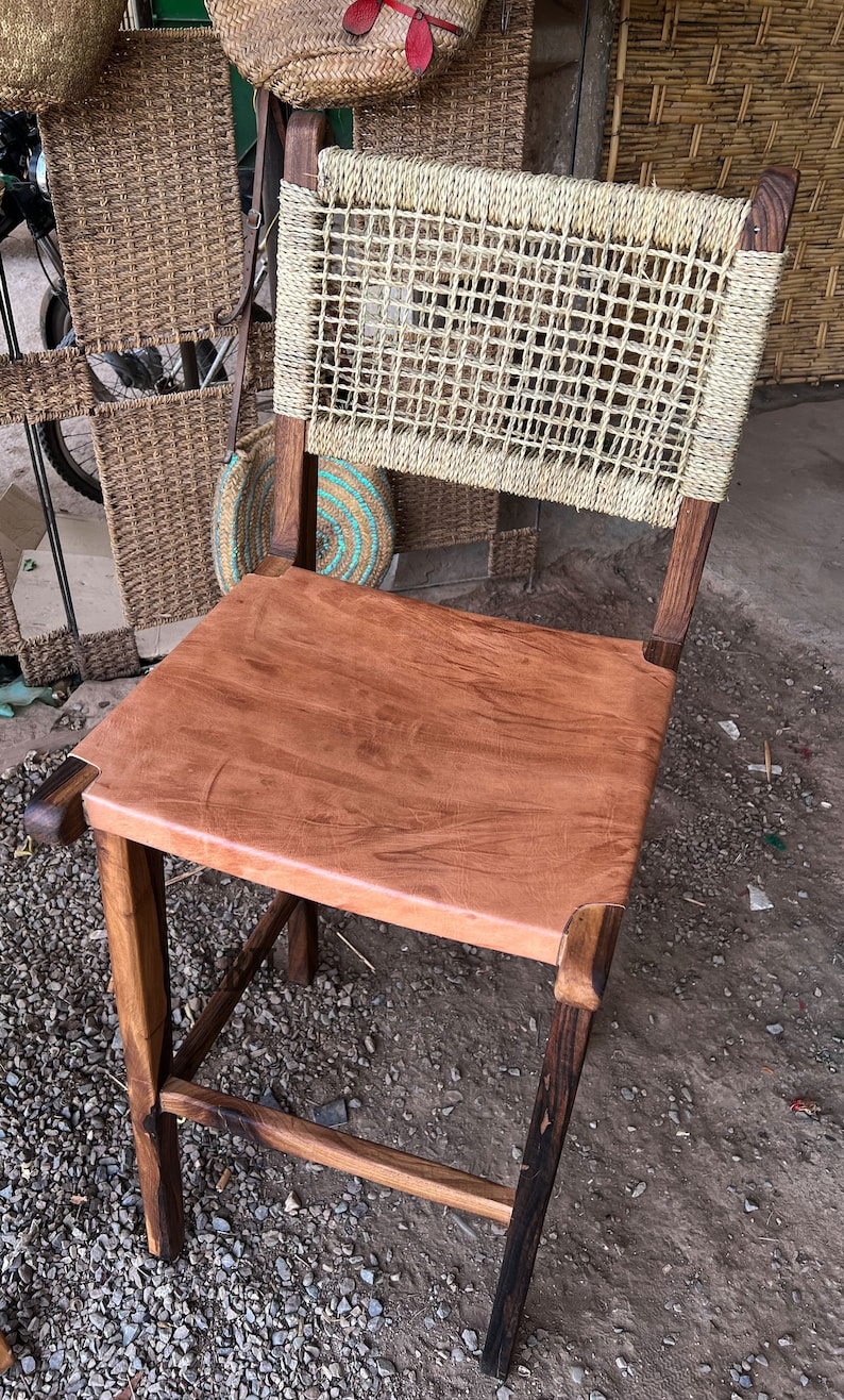 May include: A wooden chair with a woven back and a brown leather seat. The chair has a dark brown finish and is sitting on a gravel surface.