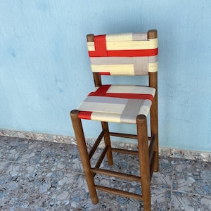 Könnte beinhalten: Handgefertigter Barhocker aus Holz mit gewebter Sitzfläche und Rückenlehne. Sitzfläche und Rückenlehne weisen ein Muster aus rotem, cremefarbenem und grauem Webmaterial auf. Der Hocker hat ein stabiles Design mit vier Beinen und einer Fußstütze. Das Holz hat eine warme, natürliche Oberfläche.