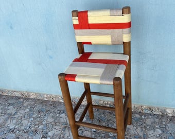 Pode incluir: Banqueta de bar de madeira artesanal com assento e encosto tecidos. O assento e o encosto apresentam um padrão de material tecido vermelho, creme e cinza. A banqueta tem um design robusto de quatro pernas com apoio para os pés. A madeira tem um acabamento quente e natural.