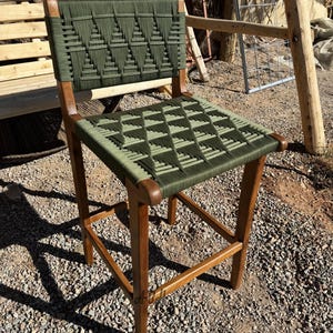 Op de afbeelding: Een houten stoel met een groene geweven zitting en rugleuning. De stoel heeft een eenvoudig ontwerp met een vierkante zitting en een licht gebogen rugleuning.