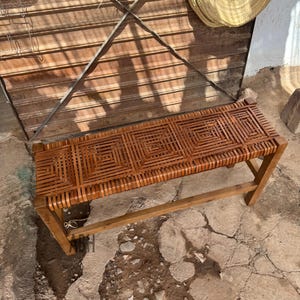 May include: A wooden bench with a woven seat. The seat is made of interwoven brown strips, forming a geometric design. The bench is rectangular and has four wooden legs. The bench measures approximately 120 cm in length.