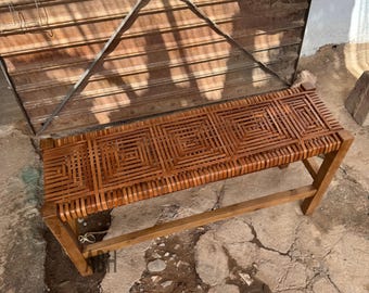 May include: A handcrafted wooden bench with a woven seat. The seat is made of interwoven strips of brown material, creating a geometric pattern. The bench has a rectangular shape and is supported by four wooden legs.