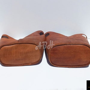 May include: Two brown leather bucket bags with stitched detailing. The bags are turned upside down, showing the bottom of the bags.