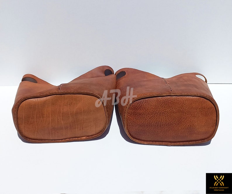 May include: Two brown leather bucket bags with stitched detailing. The bags are turned upside down, showing the bottom of the bags.